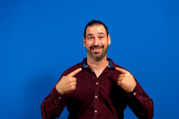 Portrait of a handsome bearded young man pointing at himself puzzled at being chosen. Amazed and attractive European man on blue background, he has a confused expression. surprise concept.