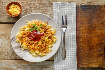 Pasta bolognese garnished with herbs and cheese in a plate on a wooden table next to a fork and a napkin.