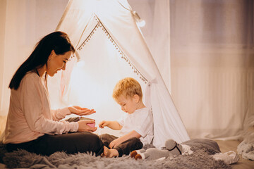 Mother with son sitting in cozy tent with lights at home on Christmas