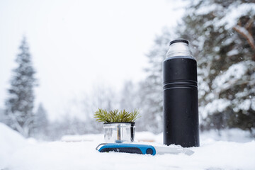 A black matte thermos, a knife and a mug with a spruce twig stand on a snow-covered stump.Warm tea in the winter frost.Minimalistic subject shot