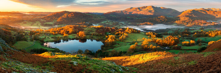 Wide panoramic view of small lake and mountains with beautiful golden sunrise light on landscape. Loughrigg Tarn, Lake District, UK. - 485892936