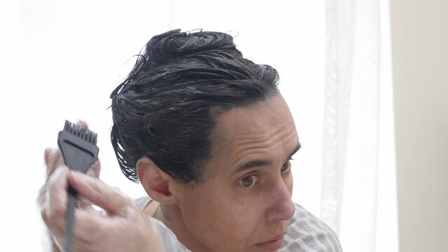 Hair coloring at home. Middle age woman dyeing hair using black brush. Woman colouring her dark hair with regrown gray roots