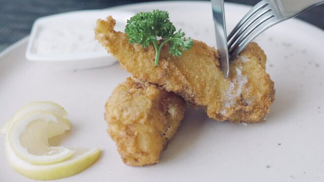 Close up 4K of knife and fork cutting delicious fish and chips on the plate with lemon and tartar sauce which is british junk food serving as appetizer for lunch. It is fillet fish and fried menu.