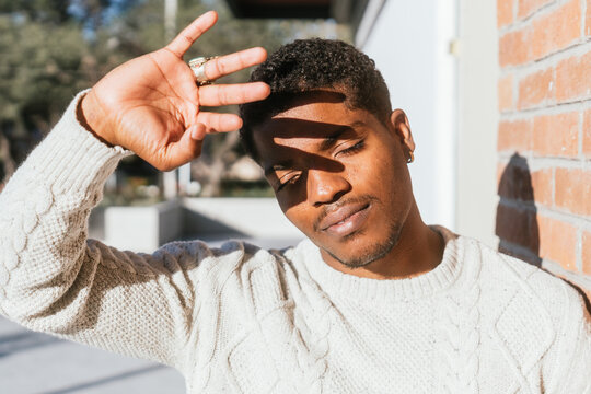 Black Man Student Covering From The Sun With His Hand. Modern Male With White Sweater Leaning Against A Wall.