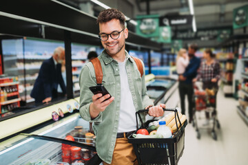 Young man buying groceries at the supermarket. Other customers in background. Consumerism concept. - 485907797