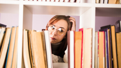 Thoughtful woman in glasses looking at books and scratching head, standing behind bookshelf. Young...