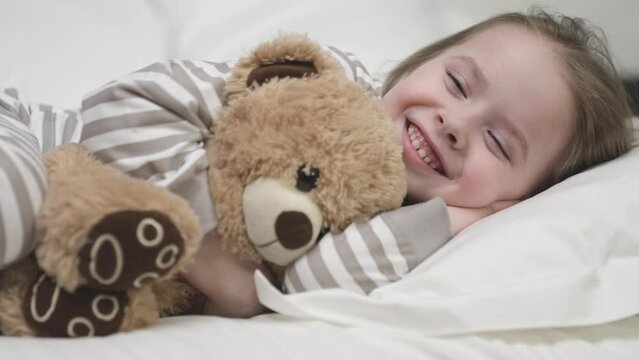 Small girl lies on pillow and smiles hugging teddy bear