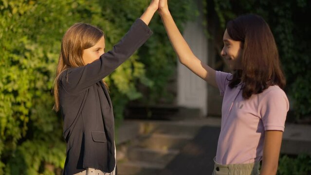 Side view two happy teenage girls gesturing high-five in slow motion at golden sunset in park. Confident Caucasian teenagers enjoying weekend leisure outdoors smiling. Friendship and lifestyle concept
