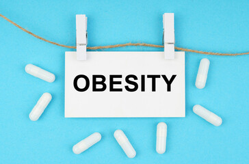 On a blue surface, on a rope with paper clips, there is a white plate with the inscription - OBESITY