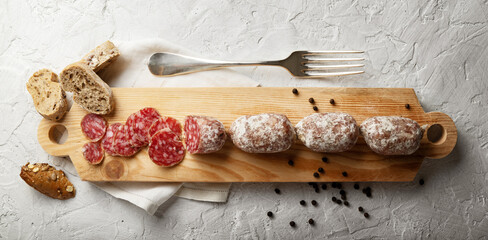 Fototapeta na wymiar Cured Italian sausages, small whole and sliced salami on wooden cutting board with bread, napkin, fork and peppercorns on white plaster background, top view, space for text.