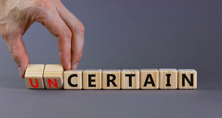 Certain or uncertain symbol. Businessman turns wooden cubes and changes the concept word uncertain to certain. Beautiful grey table, grey background, copy space. Business certain or uncertain concept.