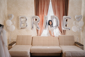 bride in white coat in a room of her house. wedding dress by sofa with balloons.