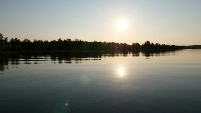 Almost completely calm lake water near the shore, the bright evening sun is reflected as in a mirror. View of an uninhabited island with the shore overgrown with small bushes, vegetation in the shade