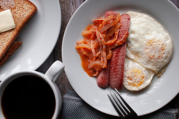 Top view, flat lay or lay flat image well done fried eggs with sausage and tomato and onion spicy salsa on a white plate with a fork on the side and a cup of black coffee with toast butter. Close up.
