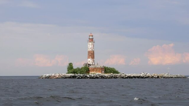 Picturesque lighthouse on tiny stony artificial island, pale colors of picture. Many sea gulls live at uninhabited islet, fly around abandoned place. Pink clouds on background