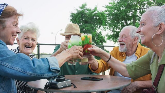 Group of happy senior friends tourists sitting outdoors in sidewalk cafe in city, enjoying drinks.