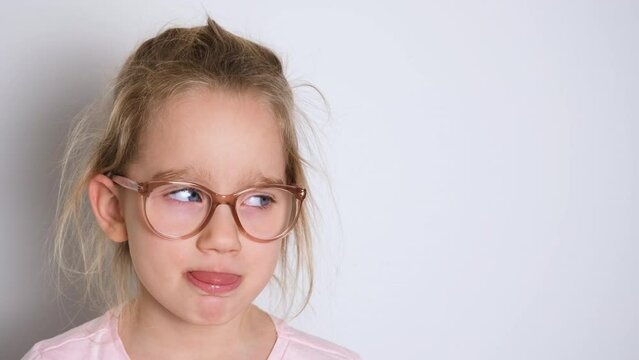 Little girl in glasses and a pink t-shirt indulges, has fun and shows her tongue, making faces at the camera. 4K resolution video banner with copy space.