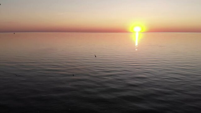 Camera flies back slowly, and shows old but working lighthouse on tiny island. Disturbed seagulls circle around, practically attacking drone. Bright sunset over the horizon, calm surface of large lake