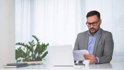 Fototapeta premium Serious focused businessman holding papers preparing rapport, analyzing business results, manager doing paperwork at workplace using computer for data analysis