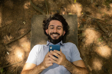 View of young man using a smartphone at day time lying down on a bench at a park. High quality photo