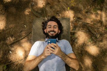 View of young man using a smartphone at day time lying down on a bench at a park. High quality photo