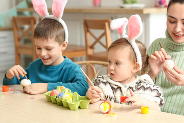 Happy little children with their mother painting Easter eggs at table