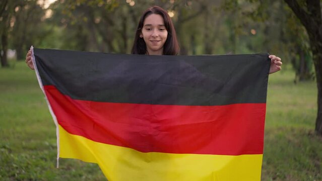 Medium shot portrait of smiling teenage girl posing with German flag in park outdoors. Happy confident teenager looking at camera standing on green meadow on summer spring day. Pride and confidence