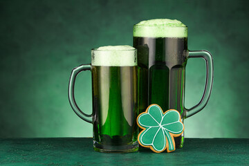 Glasses of beer and gingerbread cookie for St. Patrick's Day celebration on green background