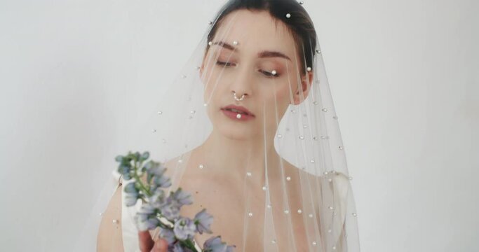 Portrait beautiful young female posing with flowers in her hands standing by clean white wall. Wedding photo session of charming sensual bride under veil. Pretty stylish caucasian woman. Bridal day
