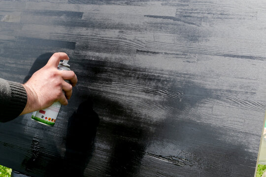 Mans male hand painting wood with black spray paint. Worker applying varnish paint on wooden table board surface, painting and caring for wood. House home renovation, copy space, texture