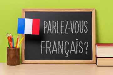 A chalkboard with the question parlez-vous francais? do you speak french? written in french, a pot with.pencils and the flag of France, on a wooden desk with green background
