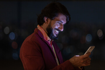 Side view of young man using a smartphone at night time with city view landscape in the background. High quality photo