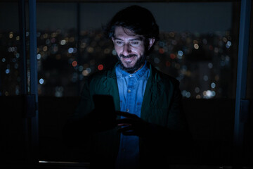Front view of young man using a smartphone at night time with city view landscape in the background. High quality photo