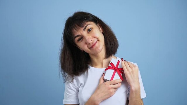 Beautiful smiling woman holding gift box in hand on blue wall background. Girl is happy with present