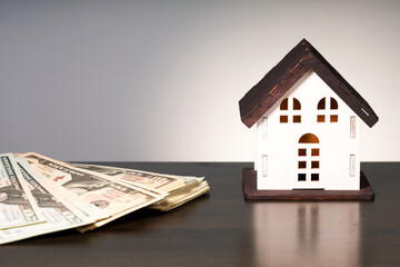 Obraz na płótnie Canvas Model of house and pile of dollar bills on desk on grey background with copy space. Real estate prices concept.