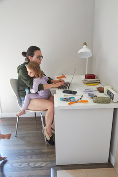 Mom Working At Home On Her Computer With A Child Sitting On Her Lap During The Pandemic