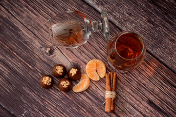 Two glasses of cognac, chocolates, a bunch of cinnamon sticks and tangerine slices on a wooden table.