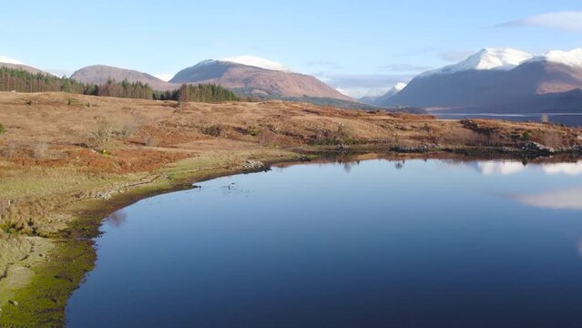 Aerial drone footage flying straight up to reveal Glen Etive and the shores of Loch Etive in the Highlands of Scotland in winter with snow-capped mountains, trees, a forest and still reflective water.