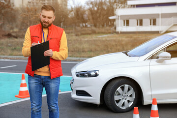 Instructor with clipboard near car on test track. Driving school