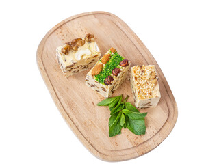 Three pieces of halva with raisins on a wooden board. Halva is decorated with walnuts, almonds, pistachios, fresh mint. White background, isolate. National sweets, organic desserts concept, top view