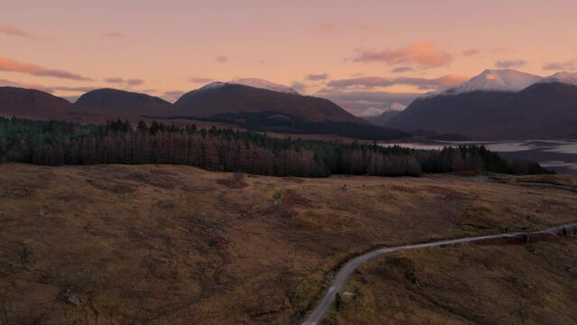 Aerial drone footage flying away from Glen Etive and Loch Etive in Scotland during an orange and pink sunrise of pastel colours, with snow-capped mountains and a forest in the background.