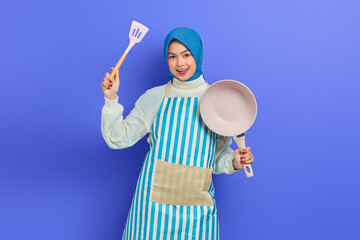 Cheerful beautiful Asian woman wearing apron holding spatula and frying pan while doing housework chores isolated on purple background. Housekeeping concept