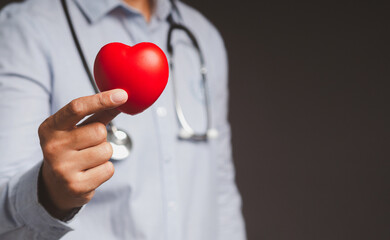 Hand of a doctor holding a red heart shape while standing on gray background in the hospital or clinic.