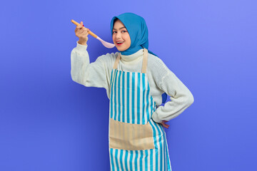Beautiful Asian woman in white sweater covered in apron and hijab holding spoon, want to taste delicious food isolated over purple background. Housekeeping concept