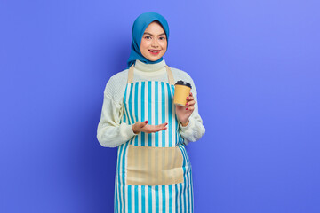 Smiling young Asian muslim woman in her 20s wearing hijab and apron showing paper coffee cup with hands isolated over purple background. People housewife muslim lifestyle concept