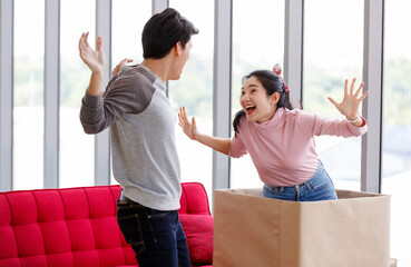 Asian young lovely lover couple female girlfriend surprise male boyfriend sitting on red sofa in living room at home by hiding inside big cardboard box as present gift for his Birthday celebration