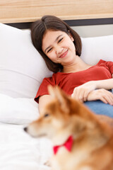 Little cute beautiful smart brown Shiba Inu dog wearing red bowtie lying aside guarding young Asian female girl owner in casual outfit laying down on bed sleeping in bedroom in blurred background