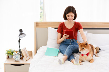 Little cute beautiful smart brown Japanese Shiba Inu dog wearing red bowtie sitting on bed together with Asian young female girl owner laying down leaning on pillow reading book and tablet in bedroom