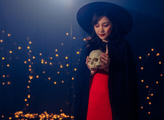 Studio shot of Asian young beautiful teenager girl wearing Halloween costume with tall witch hat black rope and orange dress standing smiling holding blue cocktail glass drink in trick & treat party