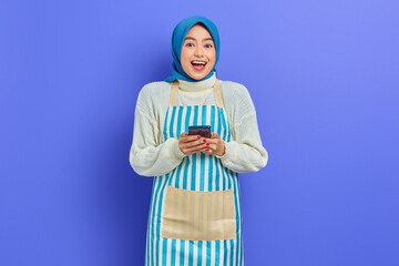 Portrait of excited young Asian muslim woman in 20s wearing hijab and apron using mobile phone while looking at camera isolated over purple background. People housewife muslim lifestyle concept